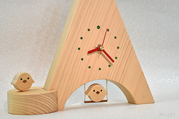 三角形に切り出した木曽檜の底辺をアーチ型に切り抜き、ブランコに乗るヒヨコをあしらった置き時計です。時計の時刻目盛りはグリーンのスワロフスキーで、12時のみ大きめのものを使用、赤い針とベースの白っぽい木曽檜でポップなイタリアンカラーに仕上げましたので、インテリアの一品として飾っても楽しいデザインです。ブランコの乗るヒヨコは軽く指で押すと、本物のブランコのように前後に揺れます。（振り子時計ではありません。）