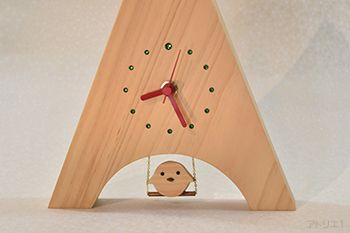三角形に切り出した木曽檜の底辺をアーチ型に切り抜き、ブランコに乗るヒヨコをあしらった置き時計です。時計の時刻目盛りはグリーンのスワロフスキーで、12時のみ大きめのものを使用、赤い針とベースの白っぽい木曽檜でポップなイタリアンカラーに仕上げましたので、インテリアの一品として飾っても楽しいデザインです。ブランコの乗るヒヨコは軽く指で押すと、本物のブランコのように前後に揺れます。