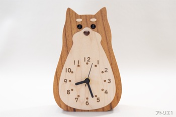 時計本体は、無垢のけやきで制作しました。白いおなかの部分はハードメープルで、時刻目盛りは柔らかい印象の数字をレーザーで刻印し、更にその内側にスモークトパーズ色のスワロフスキーを配しています。鼻はマホガニー、目玉はオニキス、麻呂眉（タン・マーキング）はハードメープルです。無垢材ならではの本物の質感と温かみがあります。